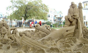 Sand sculpture of a sailor atop a dinghy that is being smashed by a shark